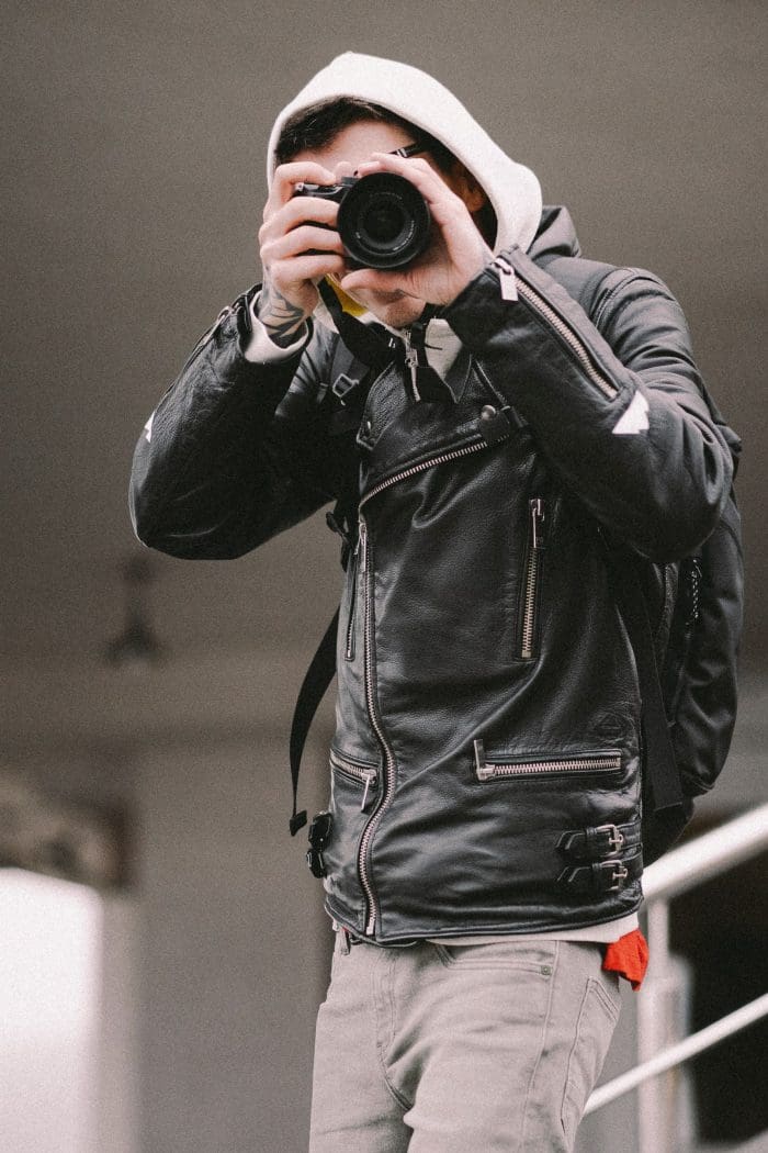 A person wearing a black leather jacket and white hoodie takes a photo with a camera, facing the lens, with a backpack on their shoulders.