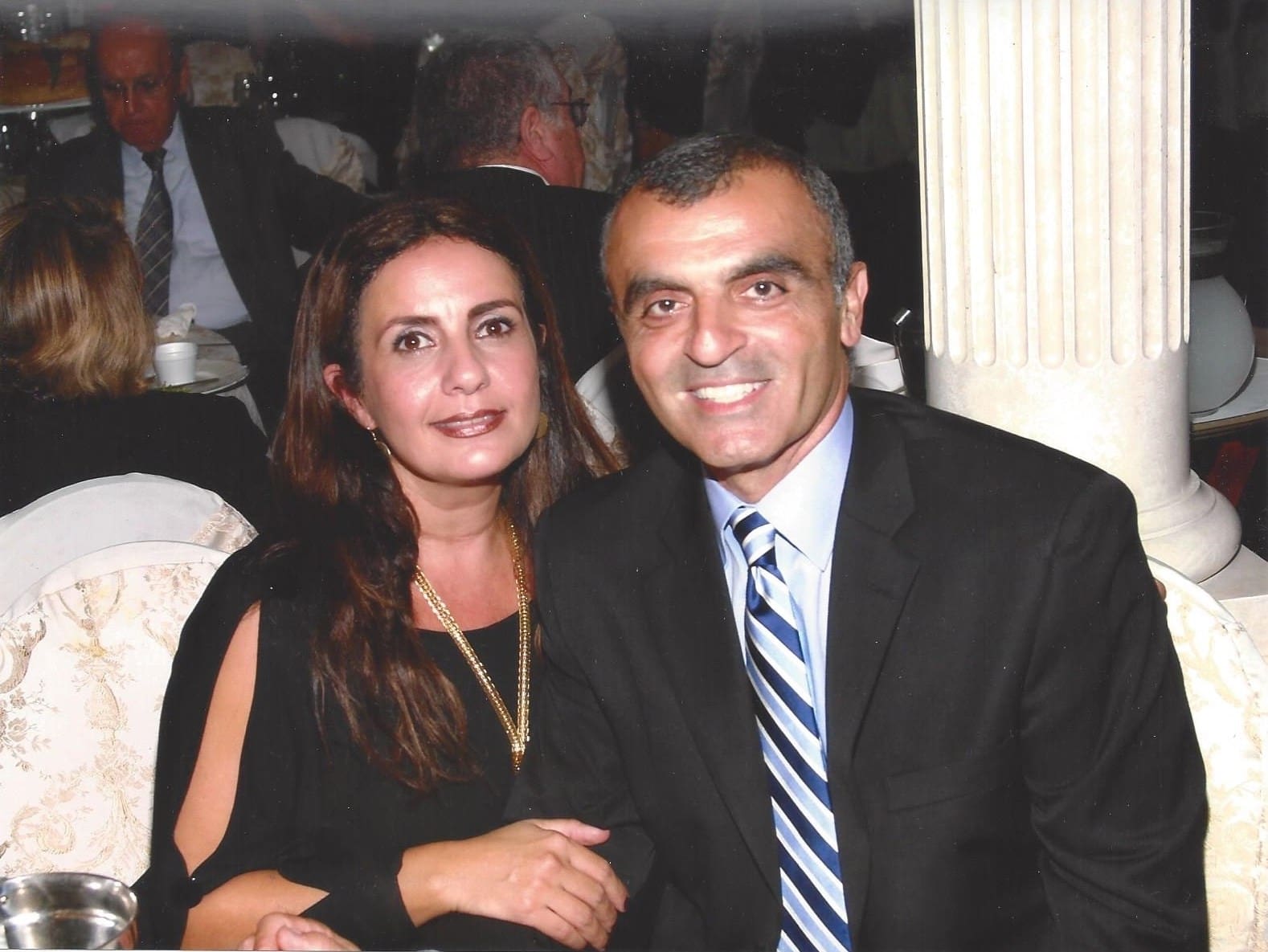 A woman and a man dressed formally are seated together, smiling at the camera in a banquet or restaurant setting with other people in the background.
