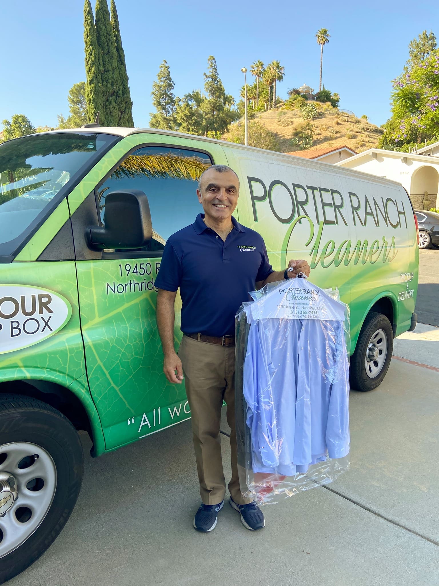 A man stands in front of a Porter Ranch Cleaners van, smiling and holding freshly cleaned, bagged shirts in a residential neighborhood.