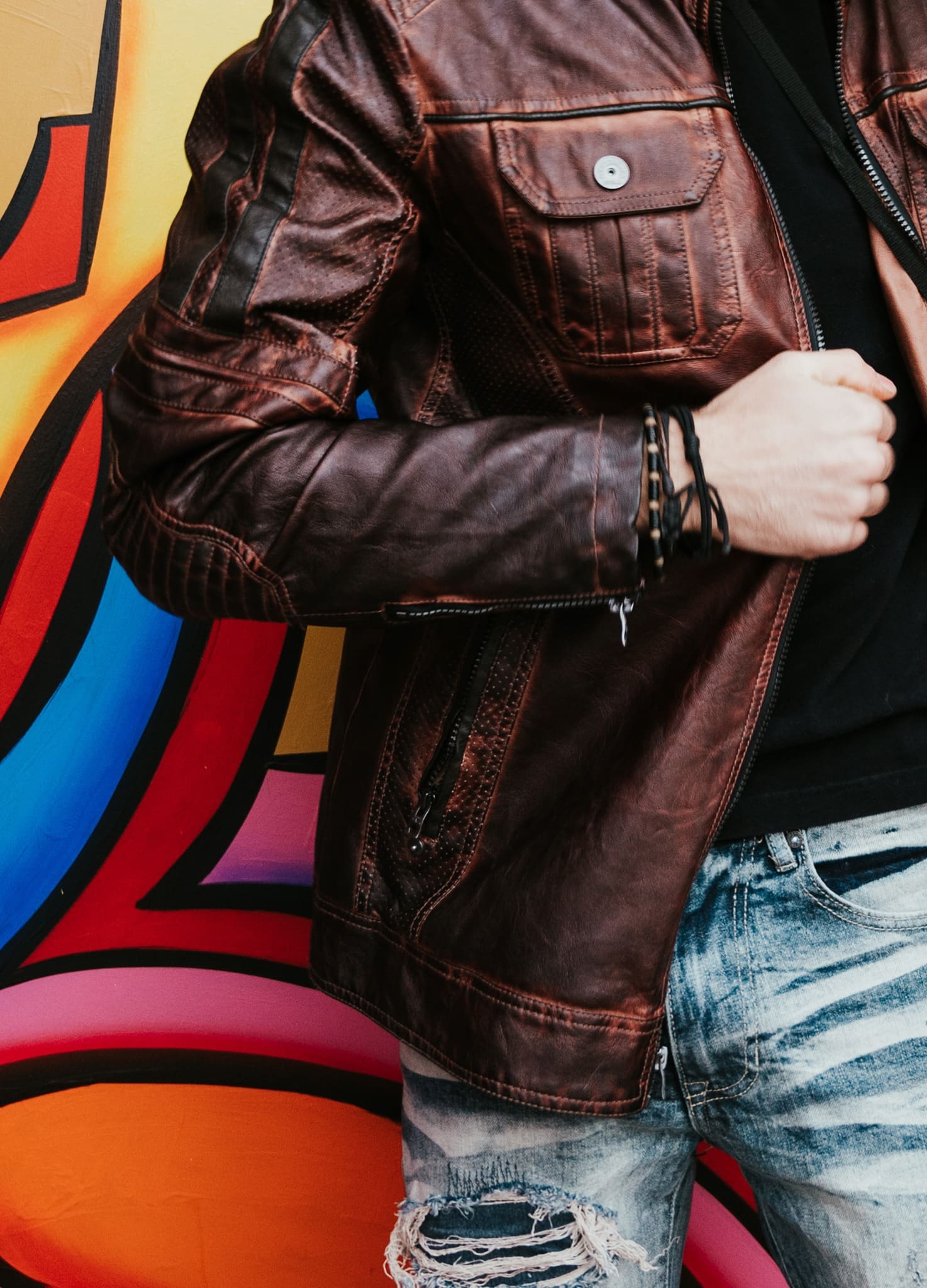 Person wearing a brown leather jacket and ripped jeans stands in front of a colorful graffiti wall.