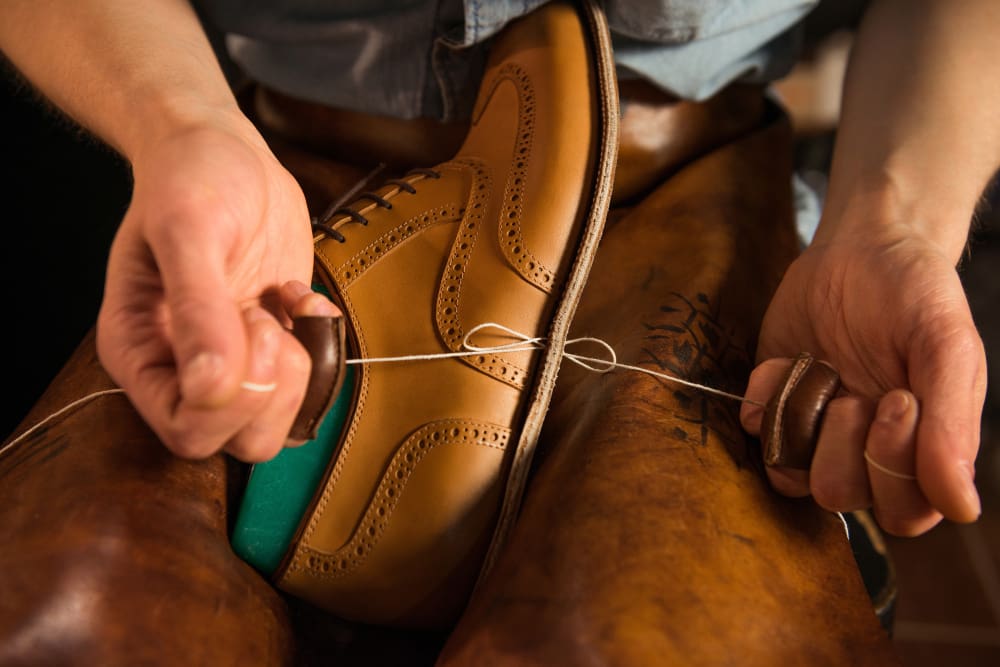 A person hand-stitching the sole of a tan leather dress shoe using thread and leather tools.