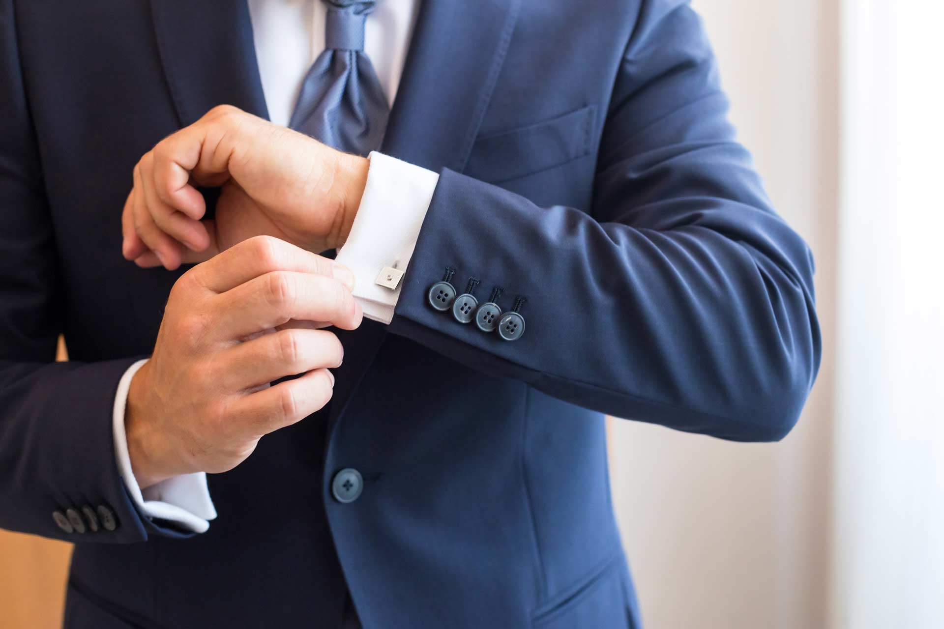 A person in a navy blue suit adjusts a silver cufflink on their white shirt sleeve, with a matching tie and buttons visible.