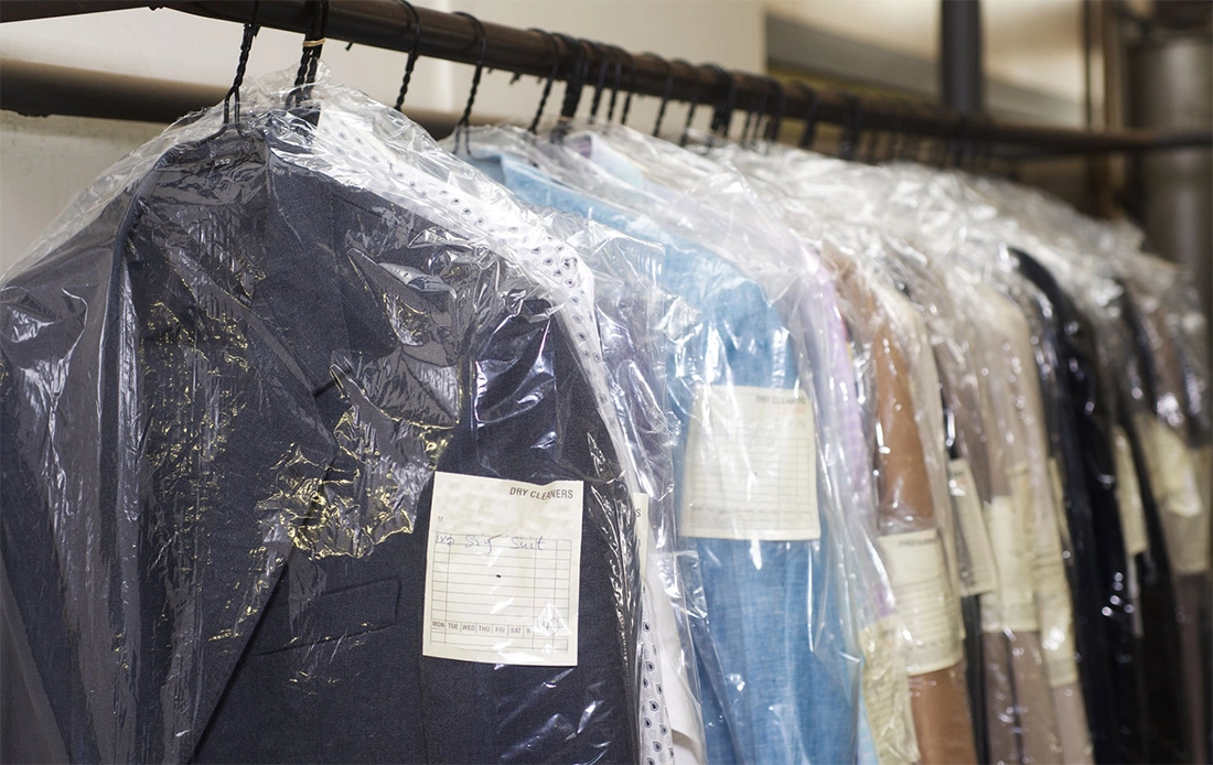 Several dry cleaned suits and shirts hang on a rack, each covered in plastic garment bags with attached paper tags.