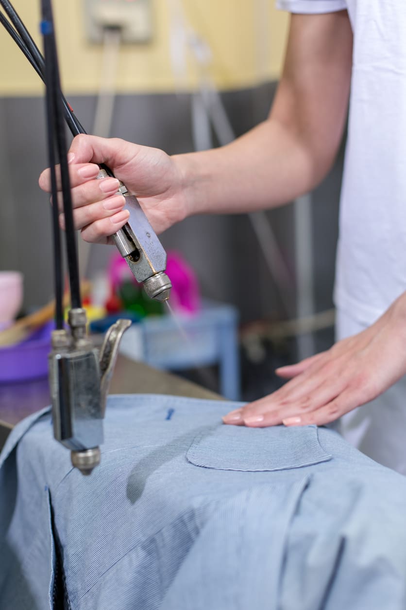 A person uses a steam iron to press a light blue shirt on an ironing board in a laundry or dry cleaning setting.
