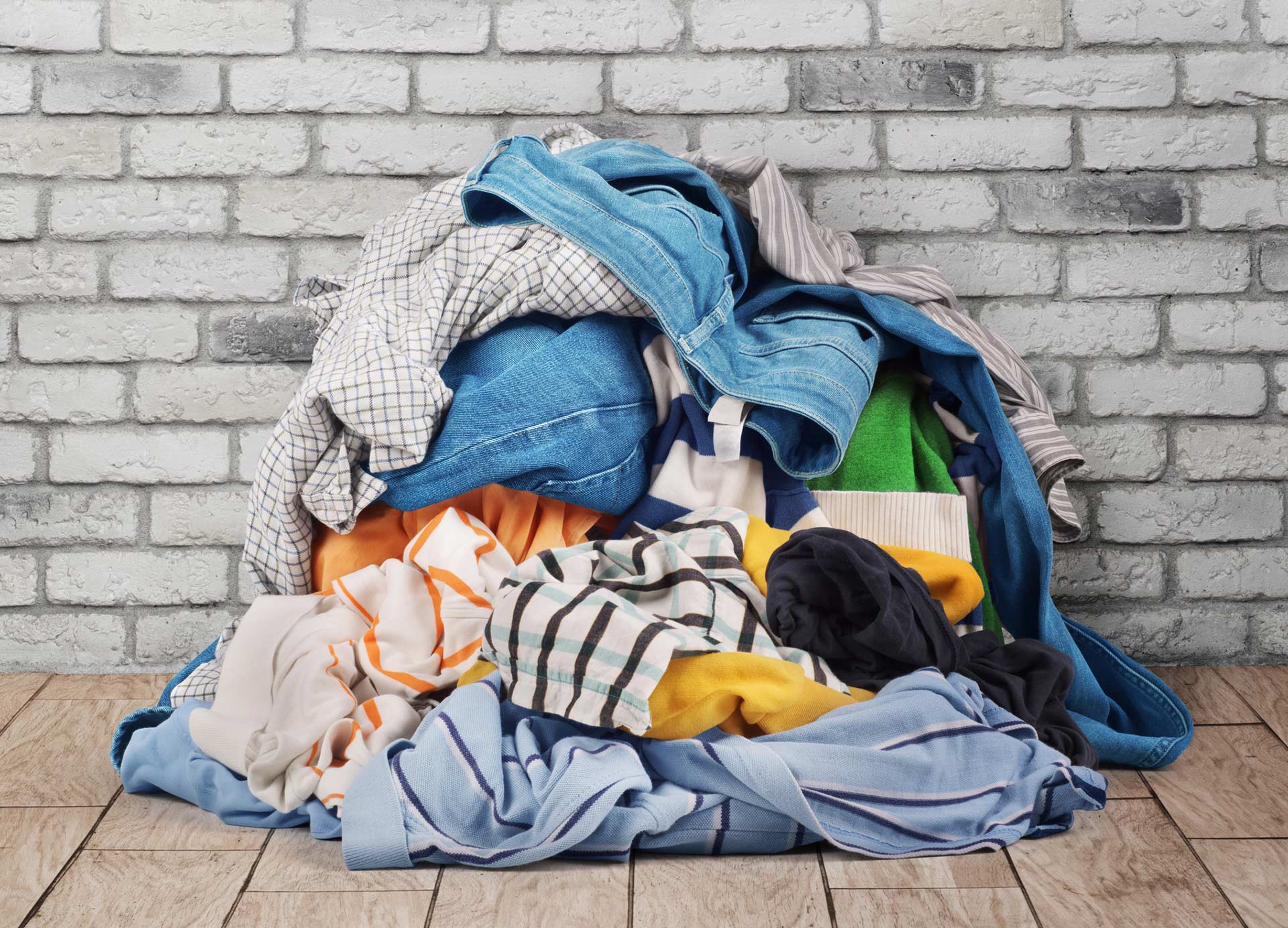 A pile of assorted wrinkled clothes in various colors and patterns is stacked on a wooden floor against a white brick wall.