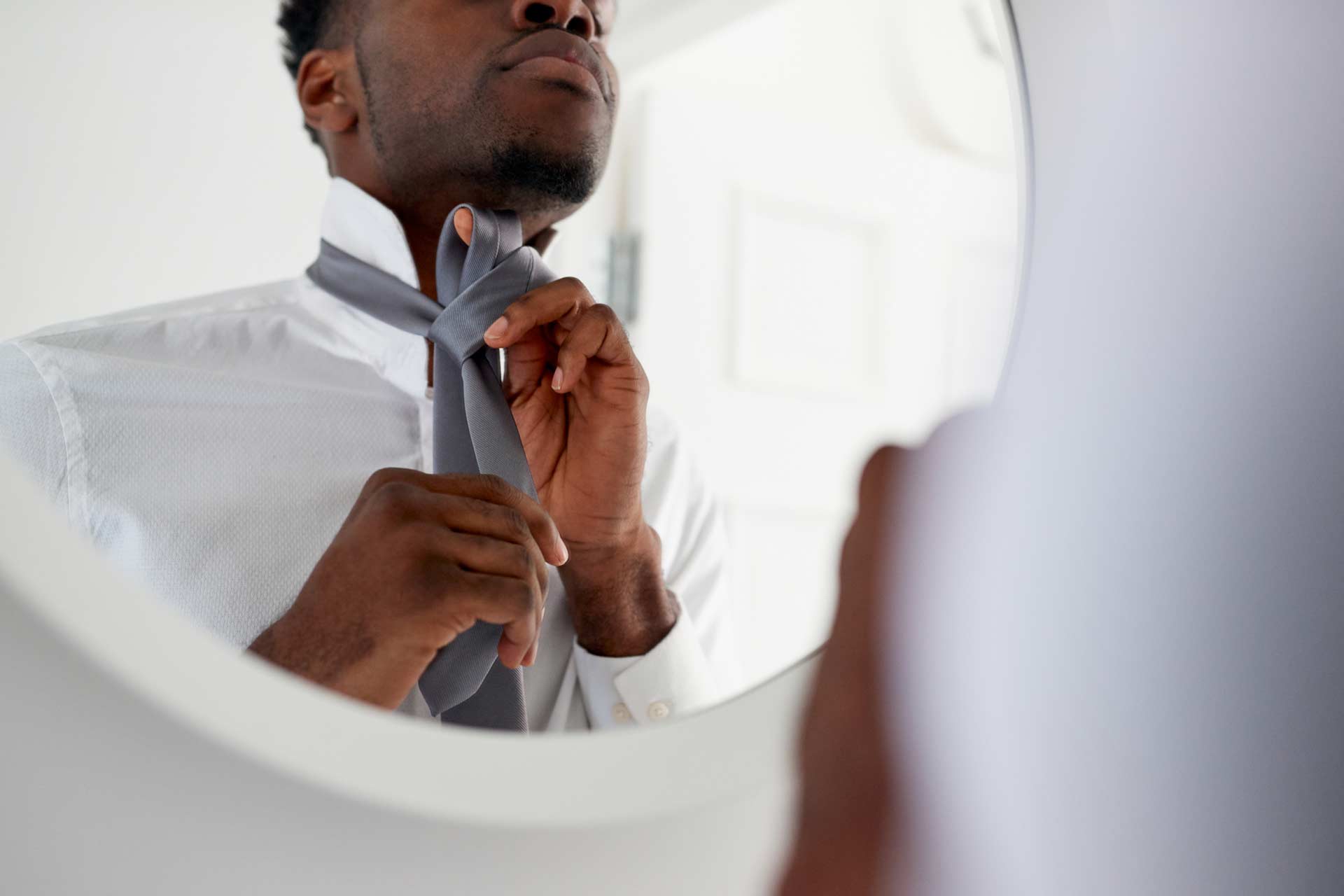 A person standing in front of a mirror ties a gray necktie while wearing a white dress shirt.