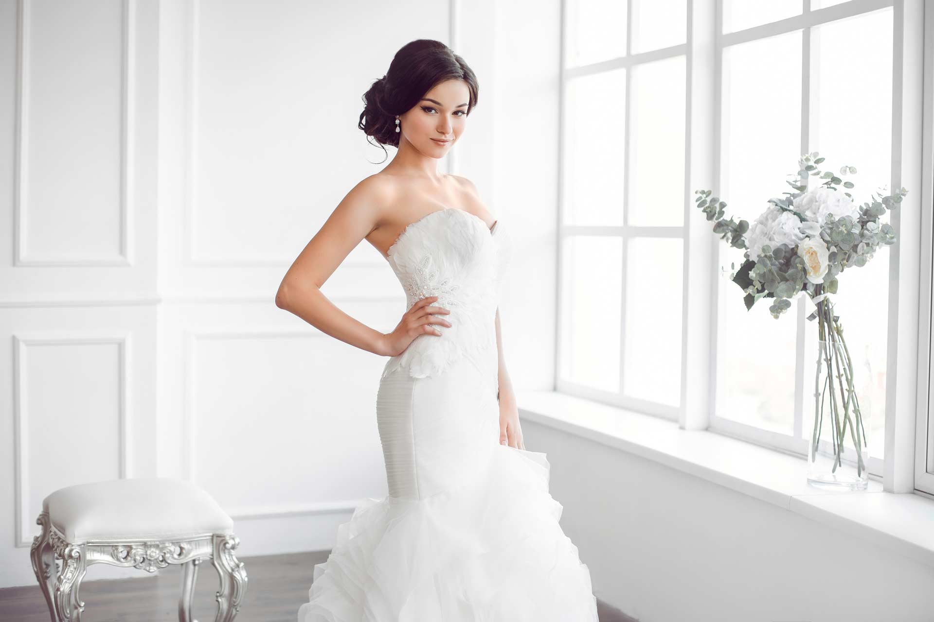 Woman in a strapless white wedding dress stands by a large window, with a white chair and a vase of flowers in the background.