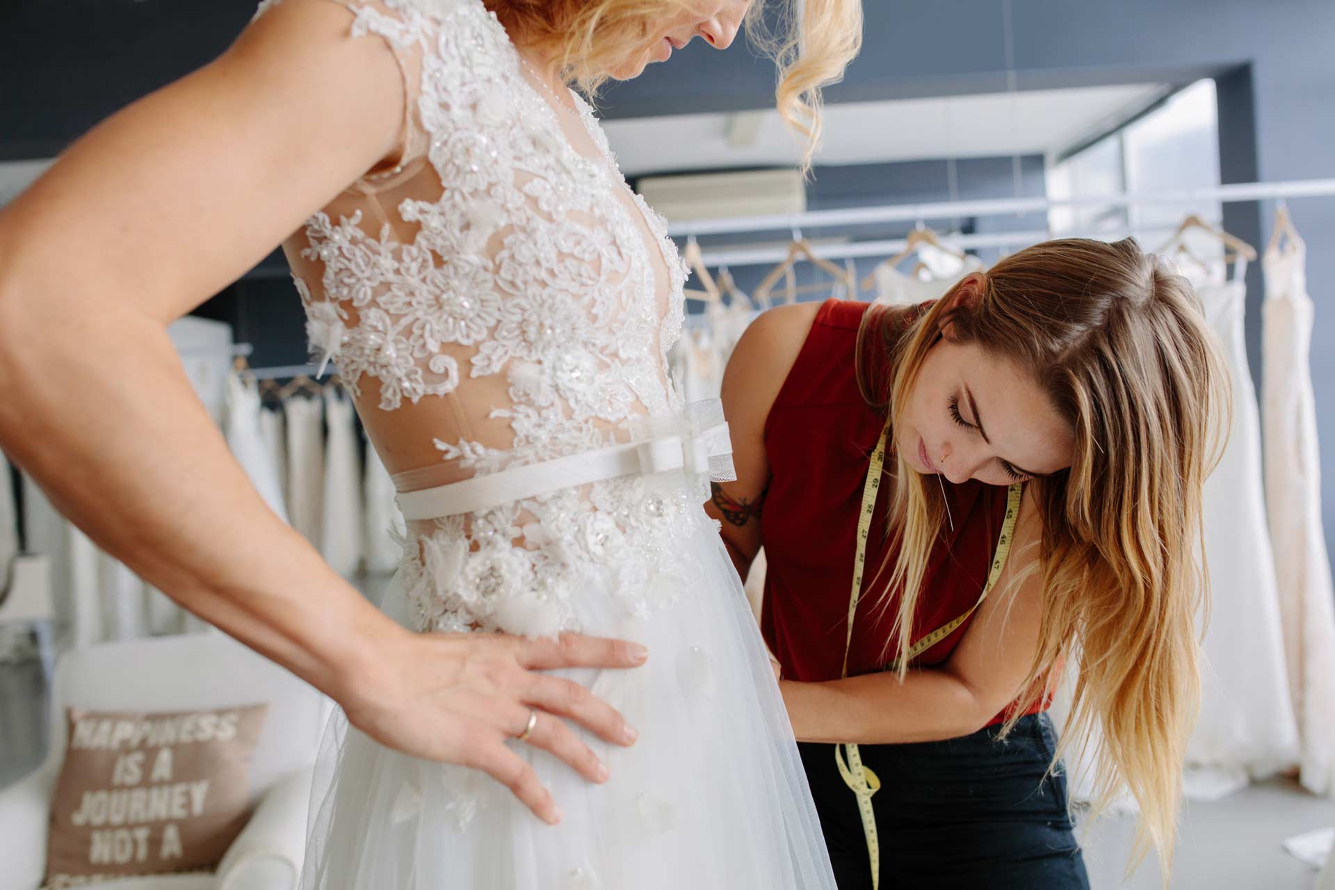 A woman wearing a white lace wedding dress is being fitted by a tailor with a measuring tape in a bridal shop.