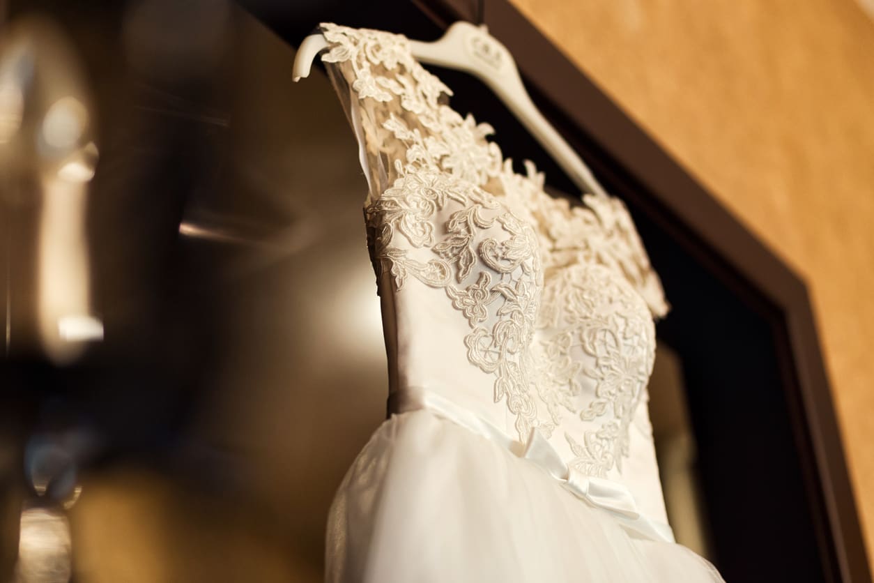 A white lace wedding dress on a hanger is displayed against a mirror, showing detailed floral embroidery on the bodice.